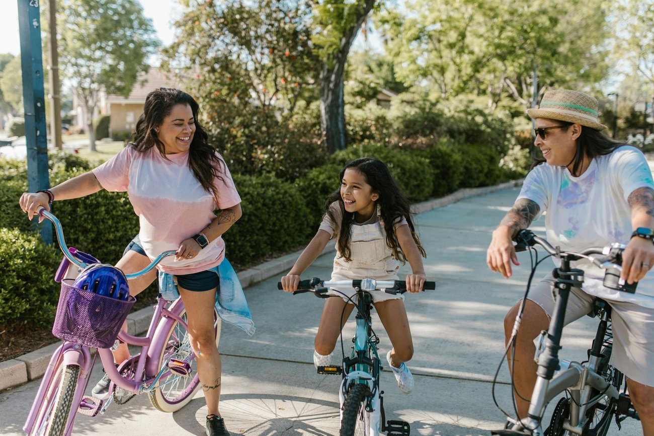 A woman, a young girl, and another woman smiling and talking while straddling bicycles on a neighborhood sidewalk.