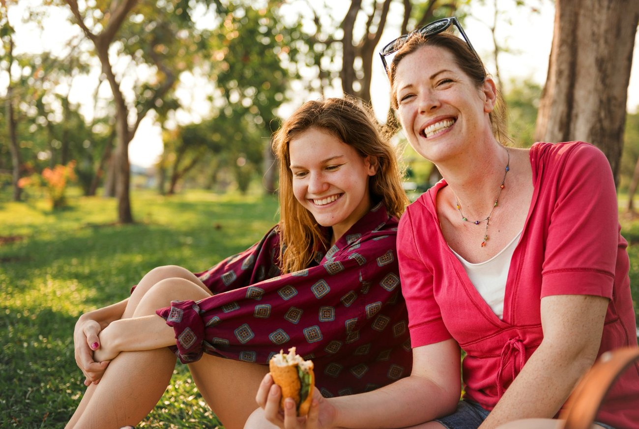 An older woman and younger woman sitting on grass, laughing together outdoors in golden afternoon light.