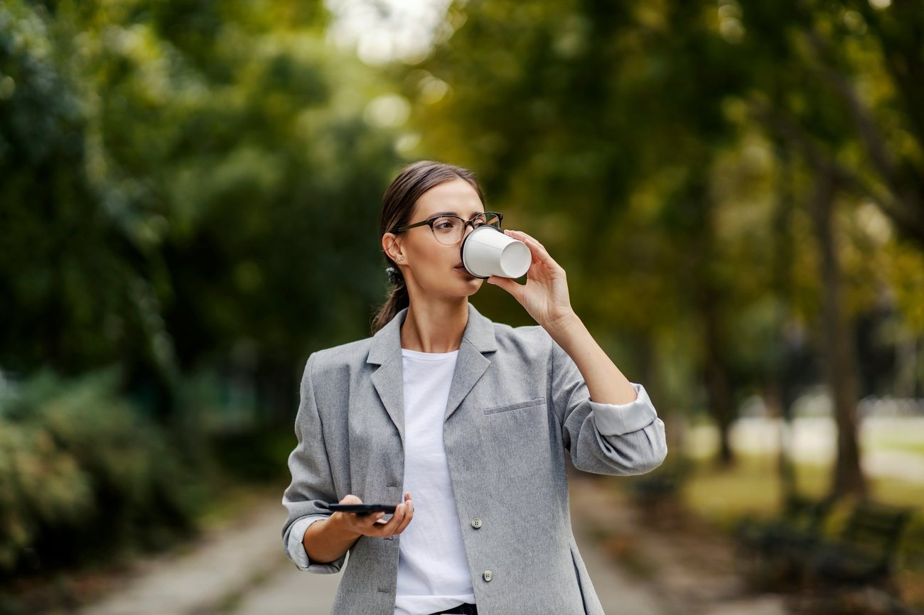 A woman in a gray blazer drinking coffee from a paper cup while holding her phone, walking outdoors along a tree-lined path.